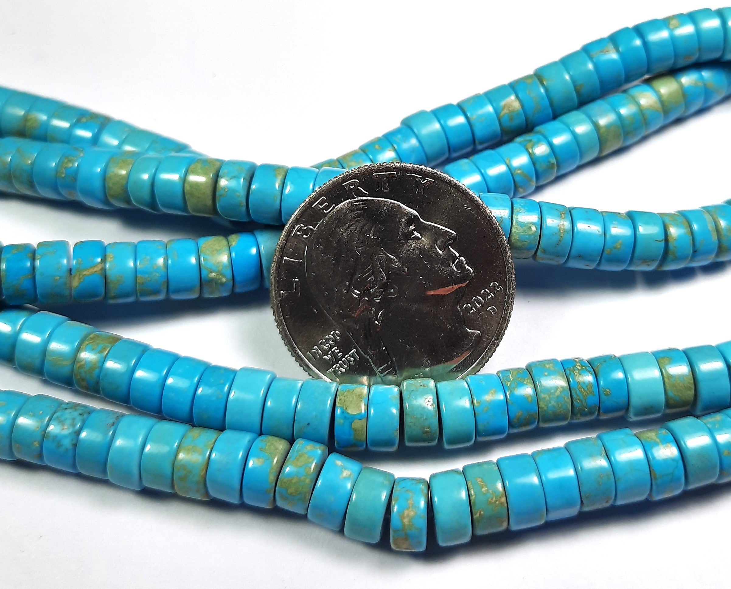 A strand of turquoise dyed white howlite beads with a flat, round shape, and a coin placed in the center for scale.